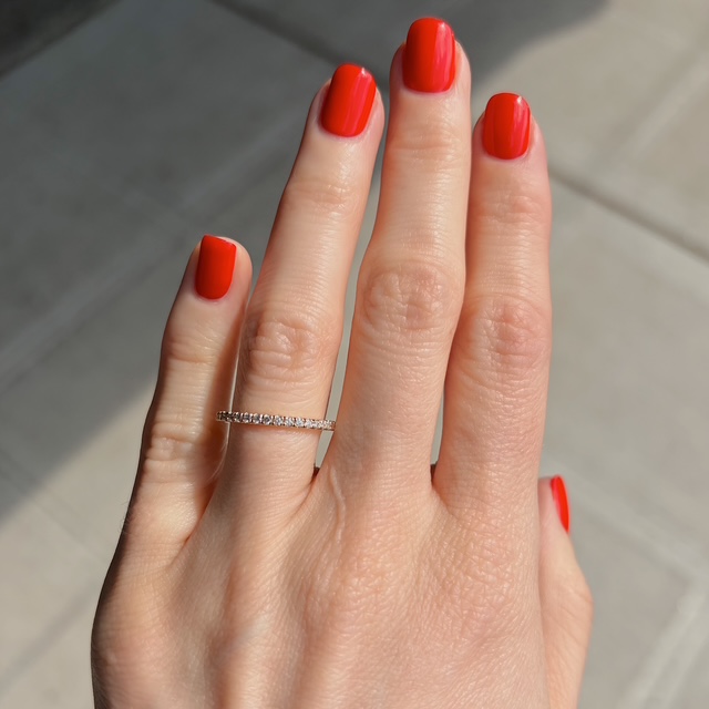 Close-up of a hand with bright red nails wearing a thin diamond eternity ring on the ring finger