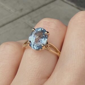 Gold ring with a large oval blue gemstone held on a finger, four-prong setting visible.