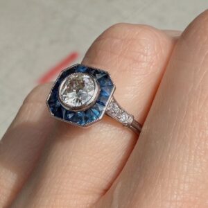 Close-up of a diamond engagement ring with a round center stone surrounded by a blue hexagonal halo on a metal band, worn on a finger.