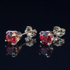 Pair of red heart-cut gemstone stud earrings on a reflective black surface, silver prong settings.