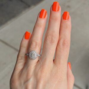Close-up of a hand with bright orange nails wearing a diamond halo engagement ring on the ring finger.