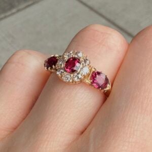 Gold ring with large red gemstone and surrounding diamonds on a finger.