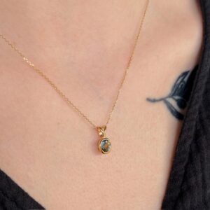 Close-up of a gold chain necklace with a small oval blue gemstone pendant resting on a person's collarbone, with black clothing and a partial tattoo visible nearby.