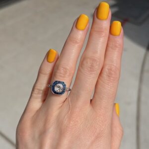 Close-up of a hand with bright yellow nail polish wearing a hexagonal blue halo ring featuring a central diamond on the ring finger.