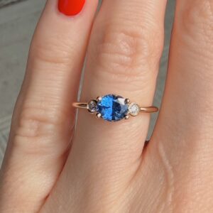 Oval blue gemstone ring set in rose gold with two small side stones on a slender band, worn on a finger with red nail polish.