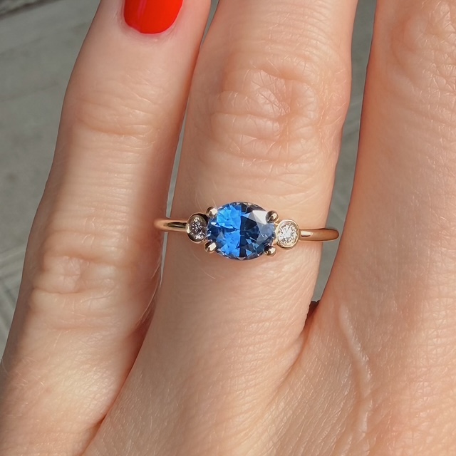 Oval blue gemstone ring set in rose gold with two small side stones on a slender band, worn on a finger with red nail polish.