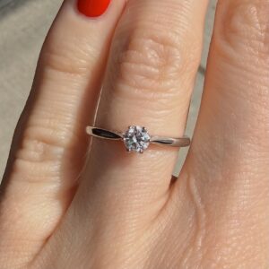 Close-up of a left hand wearing a silver engagement ring with a round-cut diamond solitaire on the ring finger, red nail polish visible.