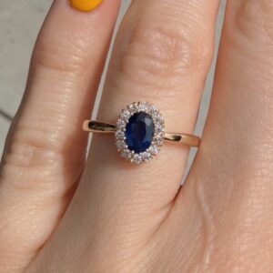Gold ring with an oval blue sapphire surrounded by small diamonds on a finger.