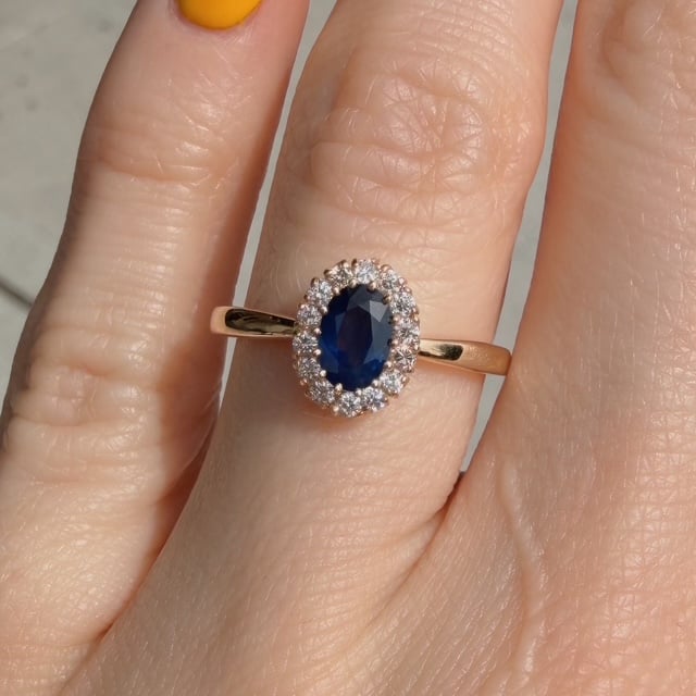 Gold ring with an oval blue sapphire surrounded by small diamonds on a finger.