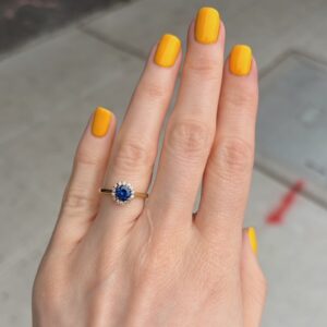 Close-up of a hand with yellow nails wearing a gold ring featuring a blue sapphire halo surrounded by small diamonds on the ring finger.