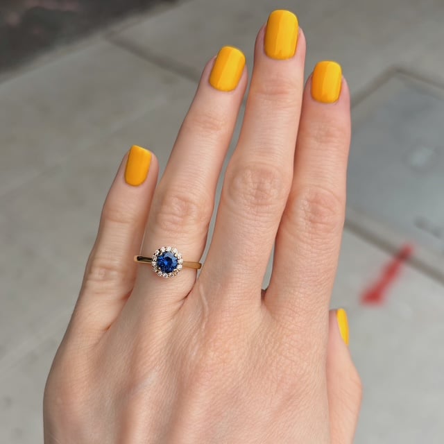 Close-up of a hand with yellow nails wearing a gold ring featuring a blue sapphire halo surrounded by small diamonds on the ring finger.