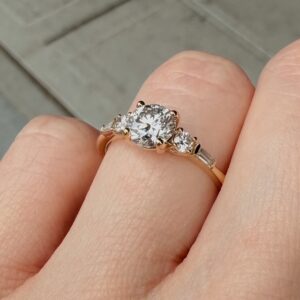 Close-up of a rose-gold engagement ring with a large round diamond center and two side stones on a finger.