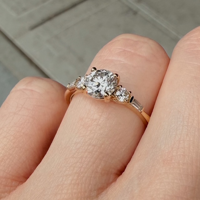 Close-up of a rose-gold engagement ring with a large round diamond center and two side stones on a finger.