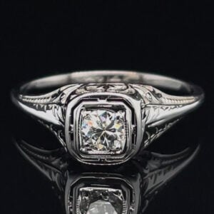 Close-up of a silver engagement ring with a square-cut diamond and engraved band on a black background.