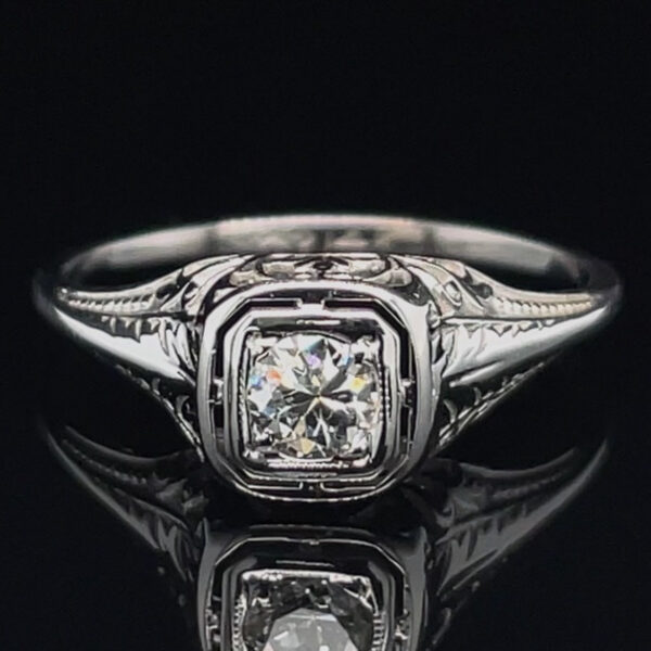 Close-up of a silver engagement ring with a square-cut diamond and engraved band on a black background.