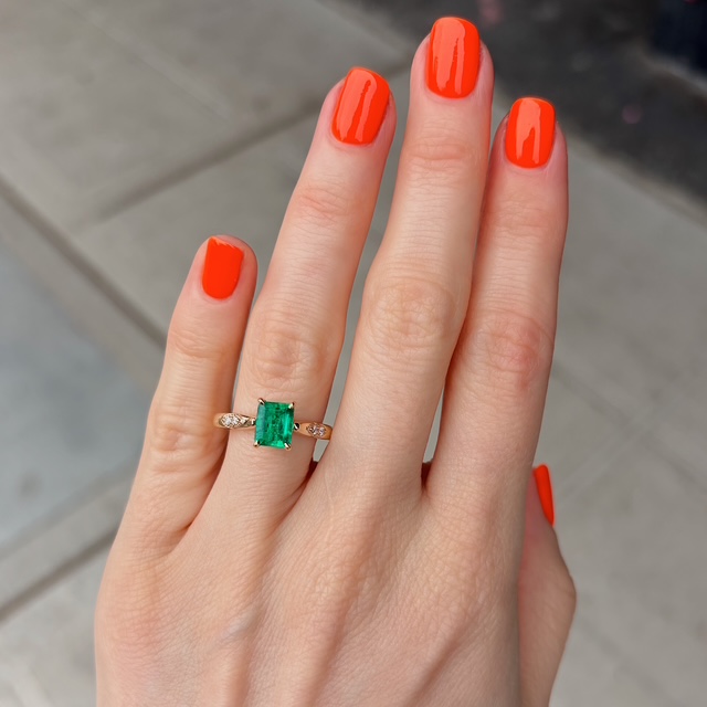 Close-up of a hand with bright orange nails wearing a gold ring with a rectangular green emerald and small diamonds on the ring finger.