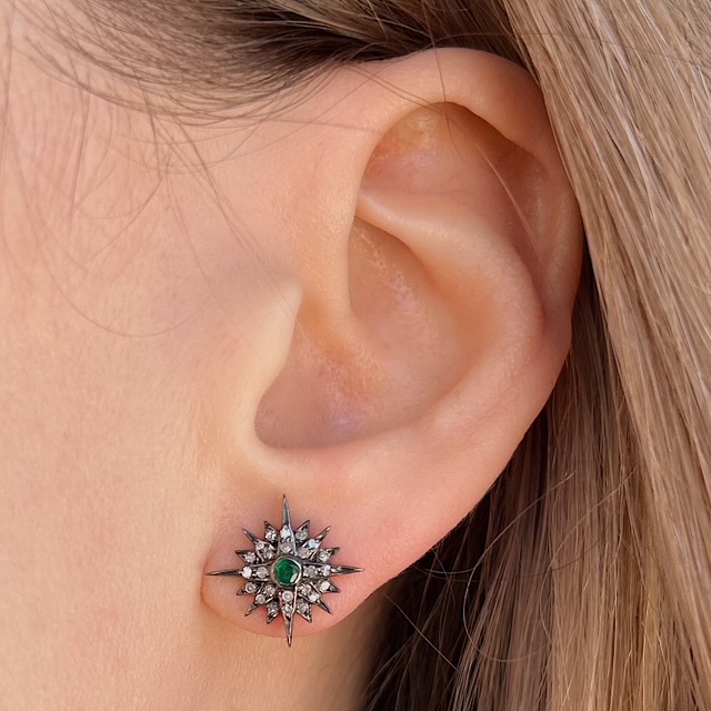 Close-up of a woman's ear wearing a silver starburst stud earring with a green central gemstone and small surrounding stones on the earlobe.