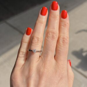 Close-up of a hand with bright red nail polish, wearing a silver solitaire engagement ring on the ring finger outdoors.