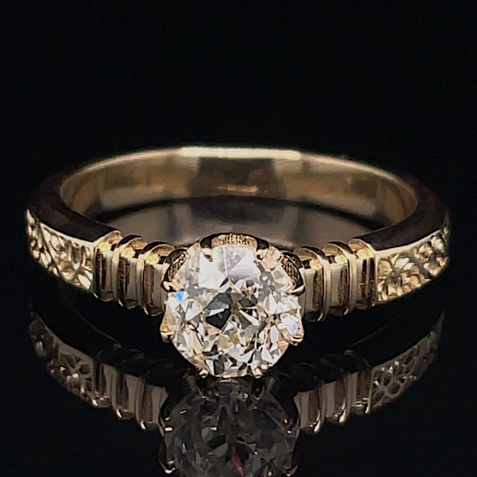 Gold engagement ring with a large round-cut diamond in a prong setting on a textured, patterned band, photographed against black.