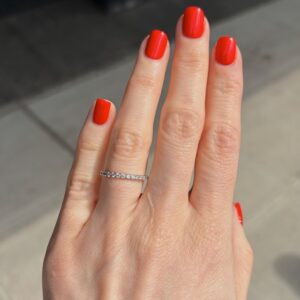 A hand with bright red nails wearing a thin diamond eternity ring on the ring finger.