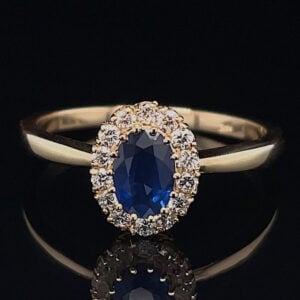 Gold ring with oval blue sapphire center surrounded by a halo of small diamonds on a black reflective surface.