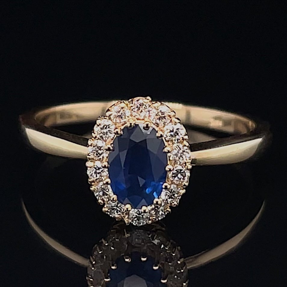 Gold ring with oval blue sapphire center surrounded by a halo of small diamonds on a black reflective surface.