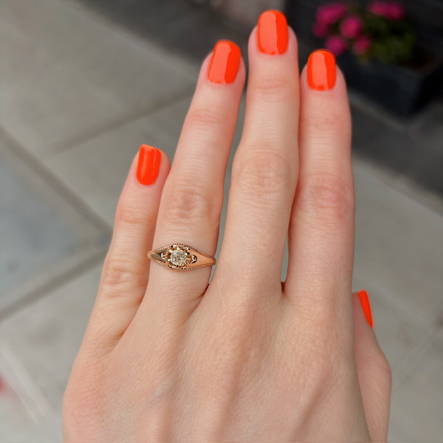 Close-up of a left hand with bright orange nails wearing a delicate gold ring with a central stone on the ring finger.