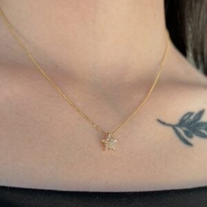 Gold chain necklace with a star-shaped pendant encrusted with stones resting on the collarbone, with a small leaf tattoo nearby on the chest edge.
