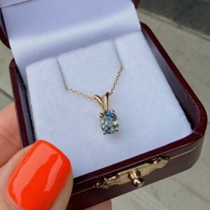 Blue oval gemstone pendant on a delicate gold chain inside an open jewelry box with a purple lining. The box sits on a curb edge, with a bright orange nail in the foreground.
