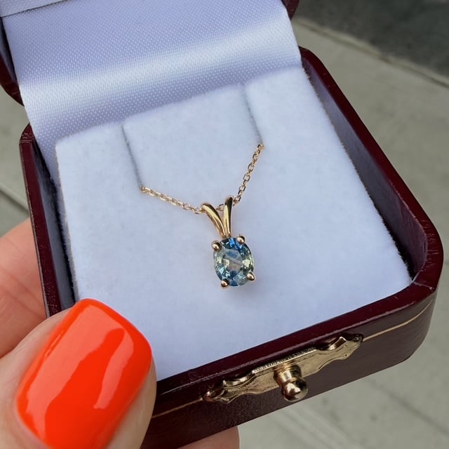 Blue oval gemstone pendant on a delicate gold chain inside an open jewelry box with a purple lining. The box sits on a curb edge, with a bright orange nail in the foreground.