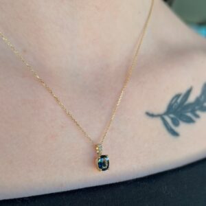 Gold chain necklace with a blue gemstone pendant resting at the collarbone, visible tattoo leaf on the chest to the right.
