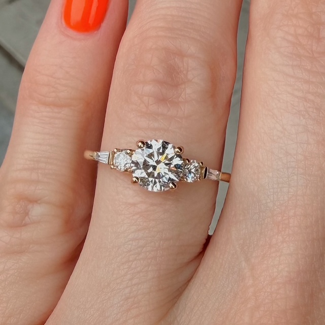 Gold engagement ring with a large central diamond and two side diamonds on a slender band, worn on the finger with orange nail polish.