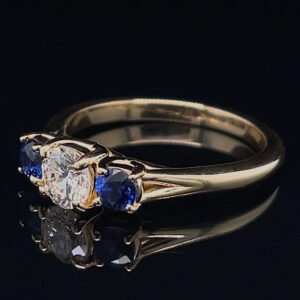 Gold ring with a central diamond flanked by blue sapphires on a black reflective surface.