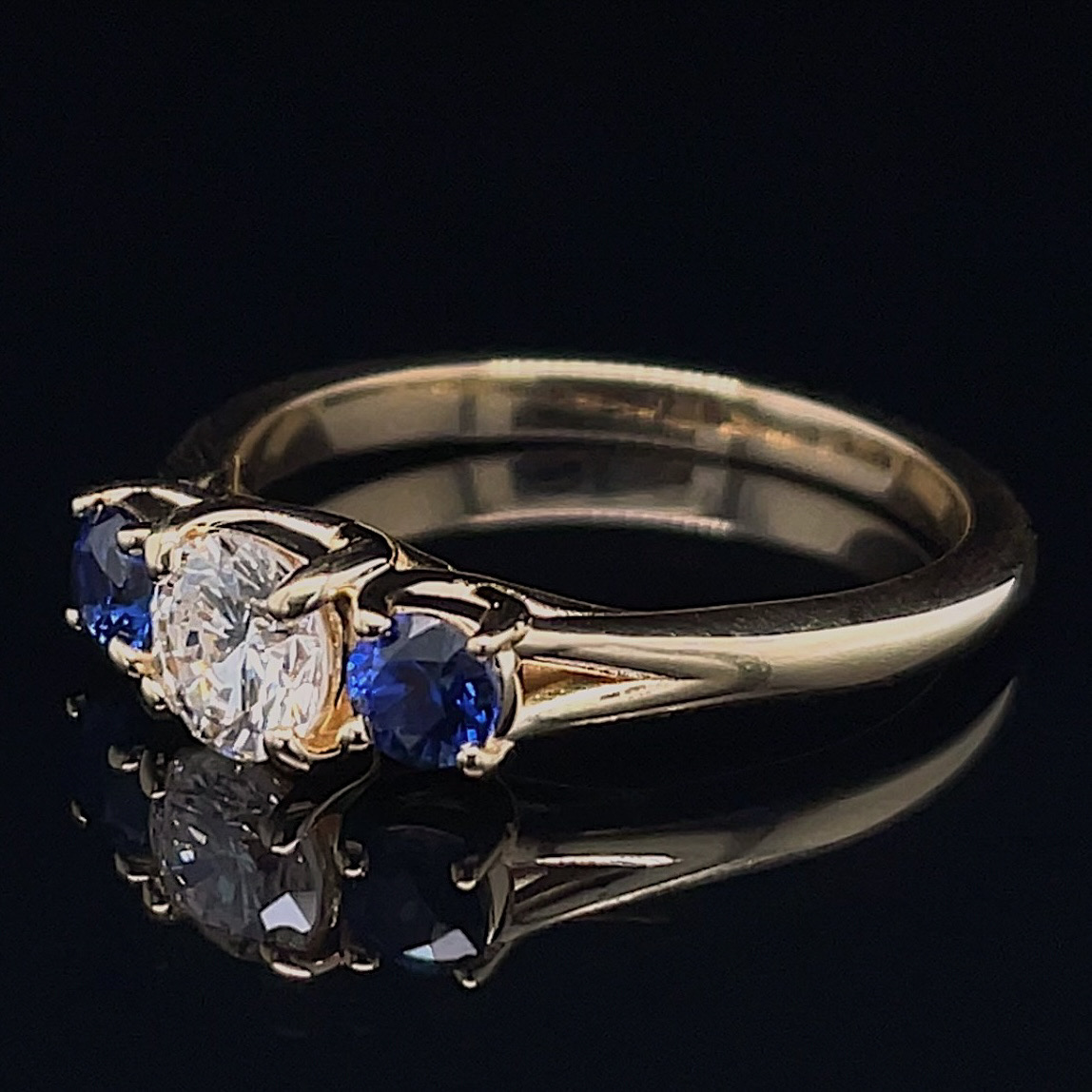 Gold ring with a central diamond flanked by blue sapphires on a black reflective surface.