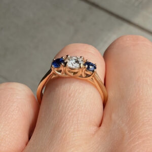 Close-up of a rose-gold ring with a central clear diamond flanked by blue sapphires on each side.