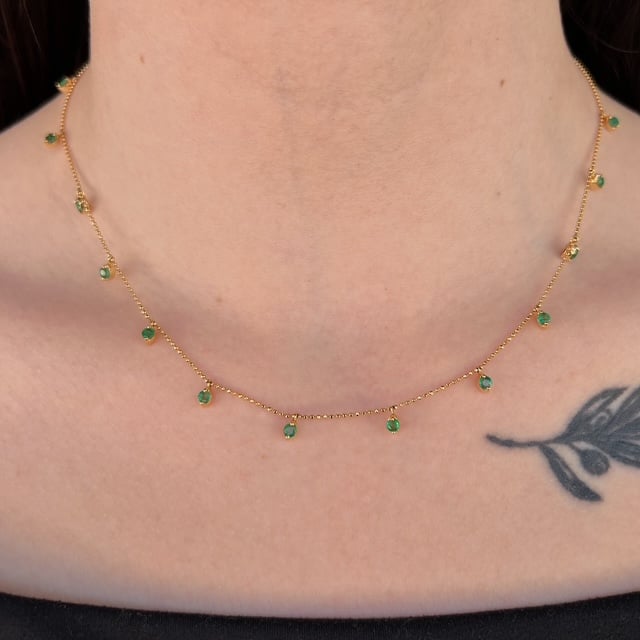Delicate gold necklace with small green stones around the neck, with part of a tattoo visible on the chest.