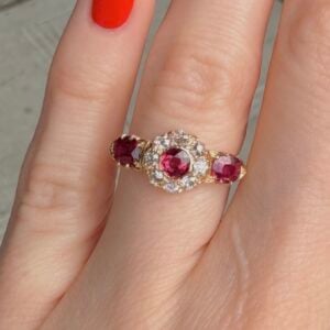 Gold ring with three pink-red gemstones and a surrounding diamond halo on a finger