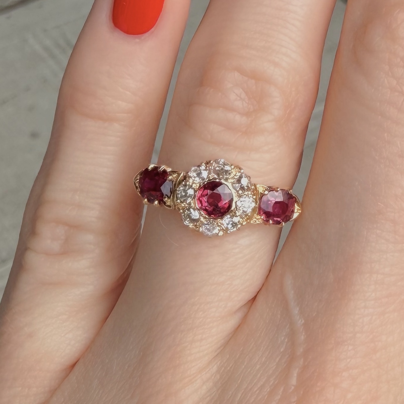 Gold ring with three pink-red gemstones and a surrounding diamond halo on a finger