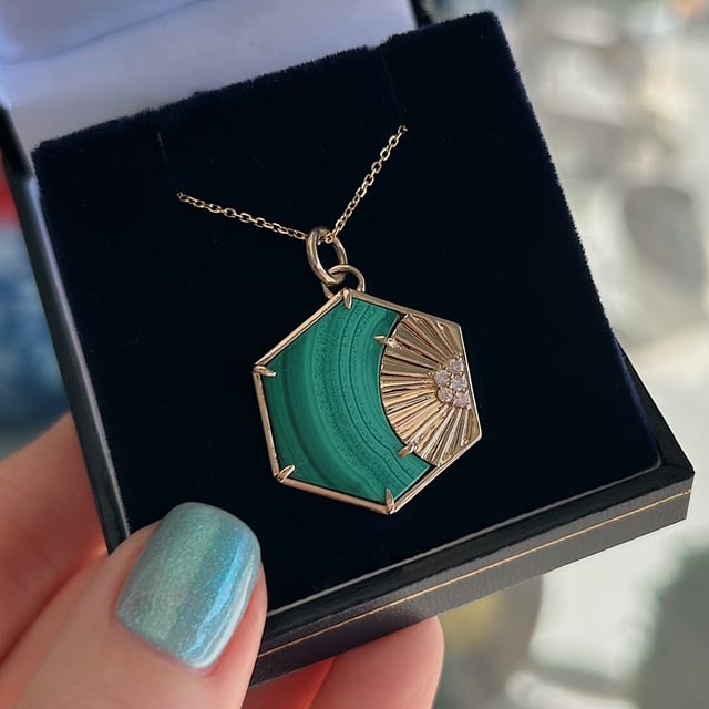 Hexagonal pendant necklace with green malachite and a gold sunburst setting, displayed in a black jewelry box.