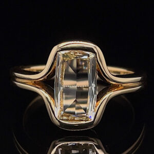 Gold ring with a rectangular emerald-cut diamond set in a wavy band against a black background.