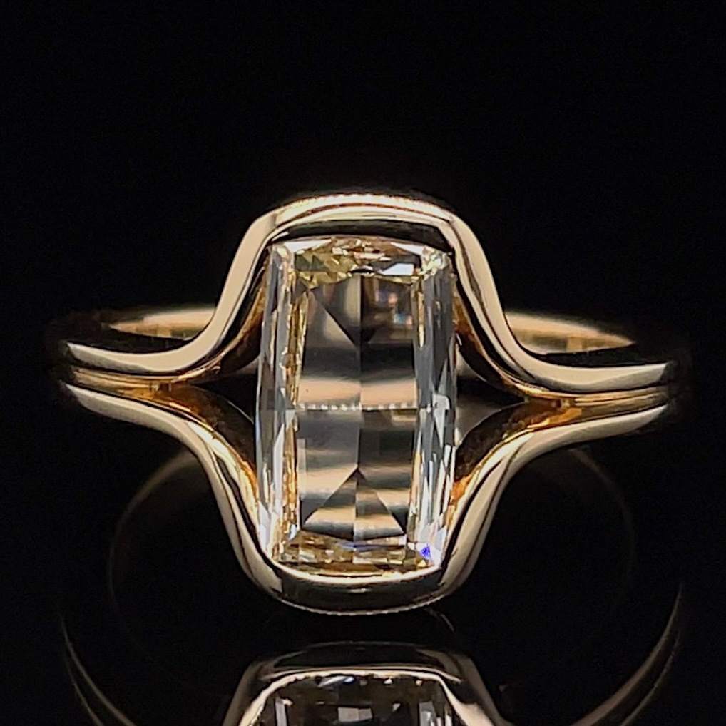 Gold ring with a rectangular emerald-cut diamond set in a wavy band against a black background.