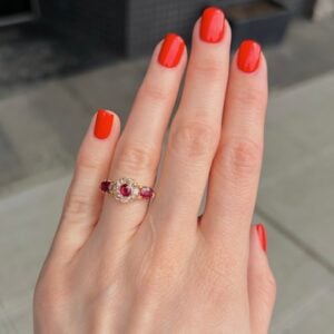 Close-up of a hand with bright red nails wearing an ornate gold ring with pink stones and small diamonds in the setting.