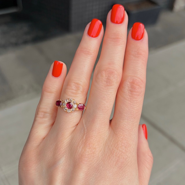 Close-up of a hand with bright red nails wearing an ornate gold ring with pink stones and small diamonds in the setting.