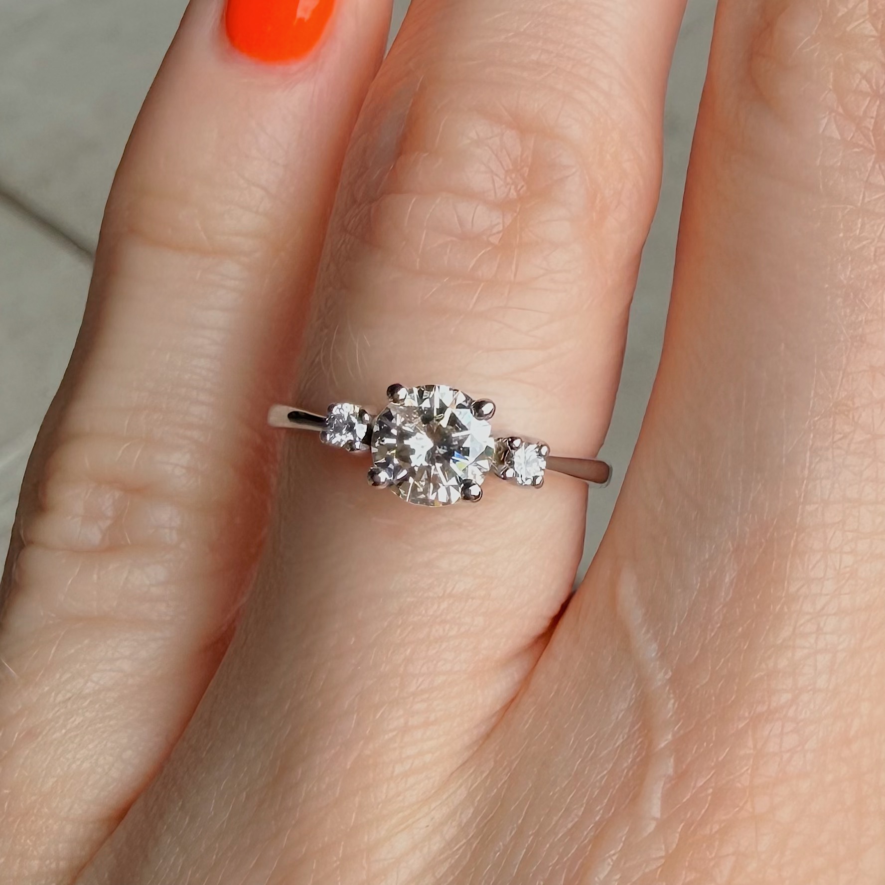 Close-up of a ring with a large central diamond and smaller side stones on the ring finger, with orange-painted nail nearby.