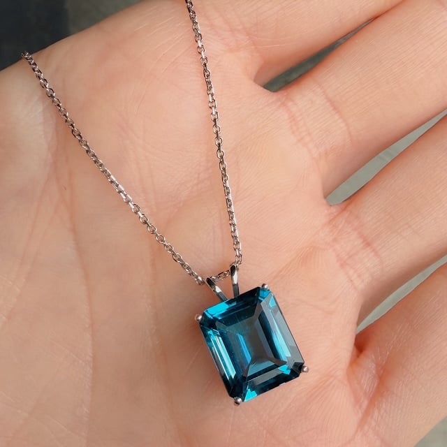Emerald-cut blue gemstone pendant on a silver chain held in an open palm.