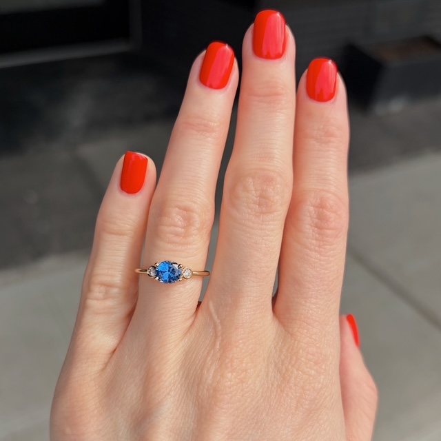 Close-up of a hand with bright red nails wearing a gold ring with a blue oval gemstone and two small side stones on the ring finger.
