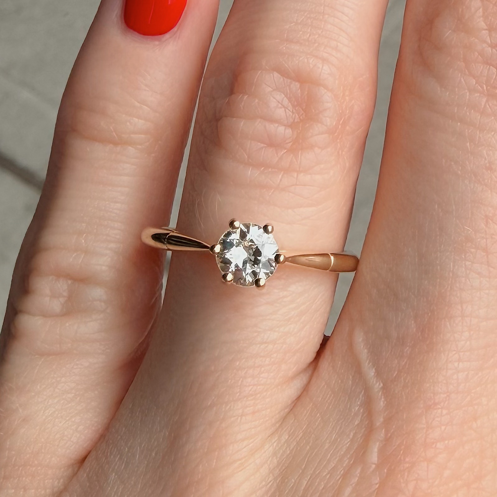 Close-up of a hand wearing a rose-gold solitaire engagement ring with a round diamond held by six prongs on a thin band, red nail polish in view.