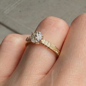 Close-up of a large round diamond engagement ring with a decorative gold leaf-pattern band on a finger.