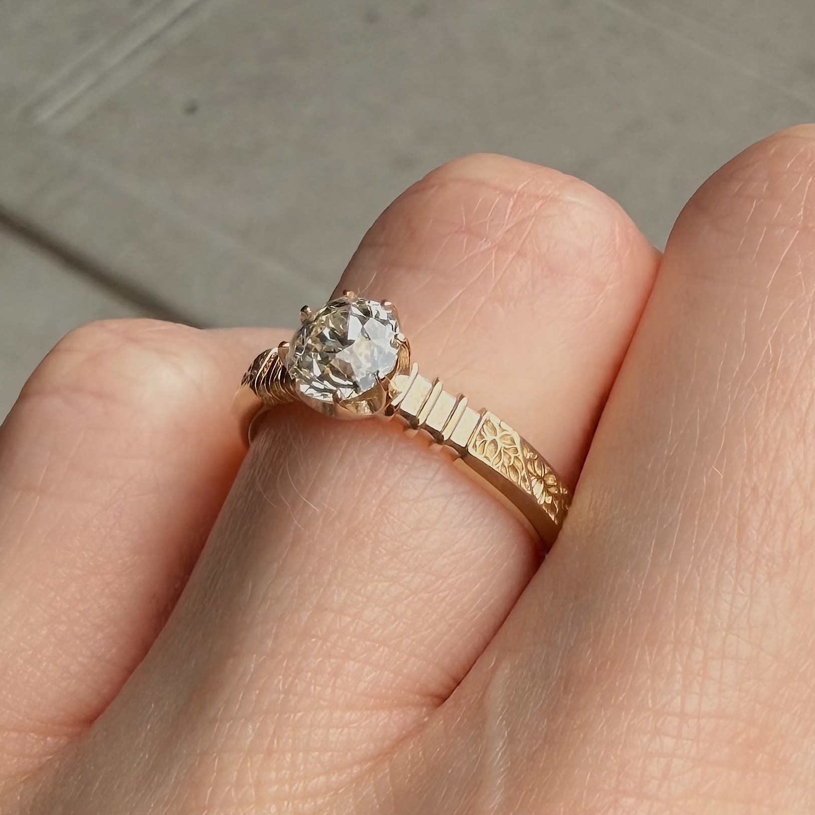 Close-up of a large round diamond engagement ring with a decorative gold leaf-pattern band on a finger.