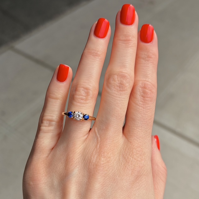 Hand with bright red nails wearing a gold ring: a central diamond flanked by two blue stones on each side.